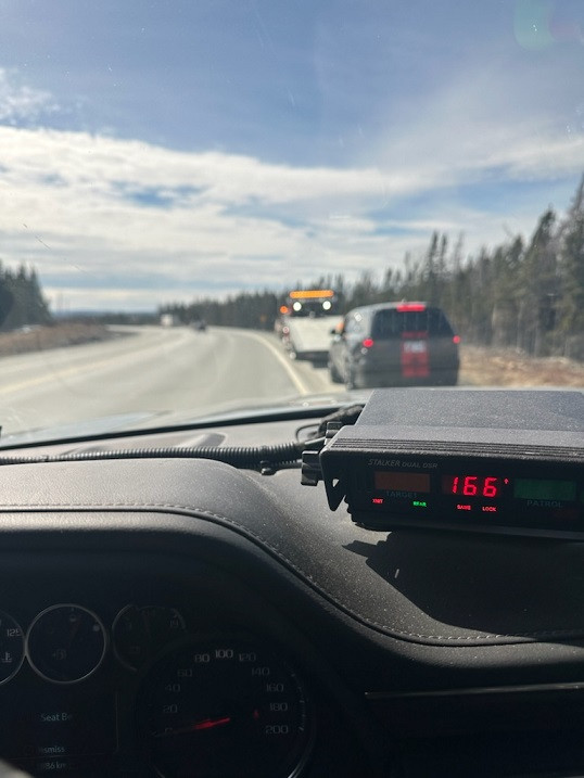A radar device mounted on the dash of a police vehicle displays a speed of 166 km/h. A vehicle and a tow truck are parked on the side of a highway in front of the police vehicle.