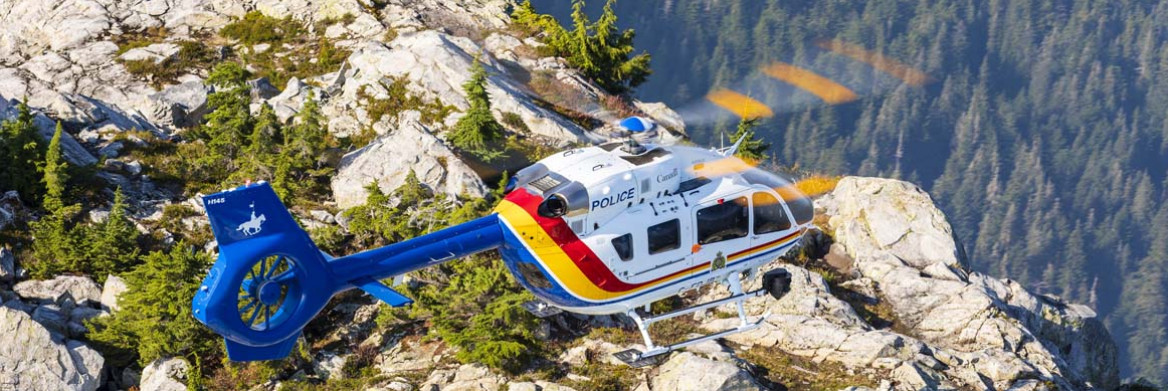 A helicopter with the word police written on it flies over a rocky mountaintop with a vast forest below. 
