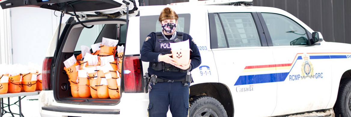 A female RCMP officer wearing a mask stands in front of a vehicle holding an orange bucket with a jack-o-lantern face. The open trunk of her vehicle is filled with dozens more buckets.