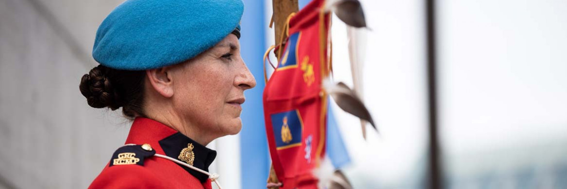 Side view of a female RCMP officer wearing red serge and a blue beret standing outdoors while holding the RCMP Eagle Staff.