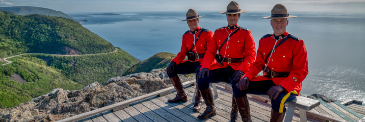 Three male RCMP officers wearing red serge sit on a bench at a lookout with a view of rolling hills and ocean.