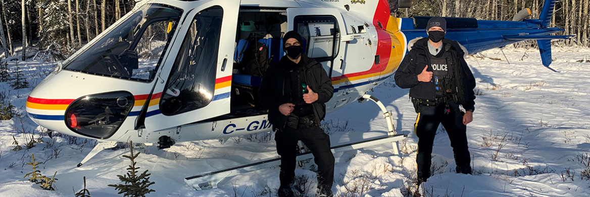 An RCMP officer and conservation officer stand in deep snow in front of a helicopter giving a thumbs up. 