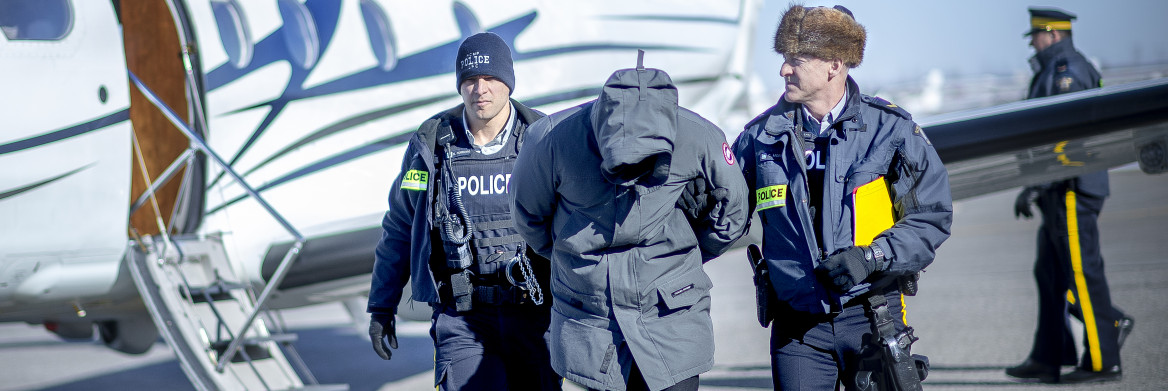 Two male RCMP officers escort a male suspect in handcuffs across an airport runway with an airplane parked in the background. 