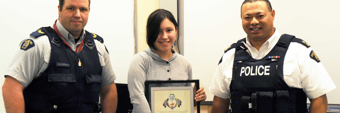Two male police officers stand next to an adolescent girl who is holding a framed image of a crest.