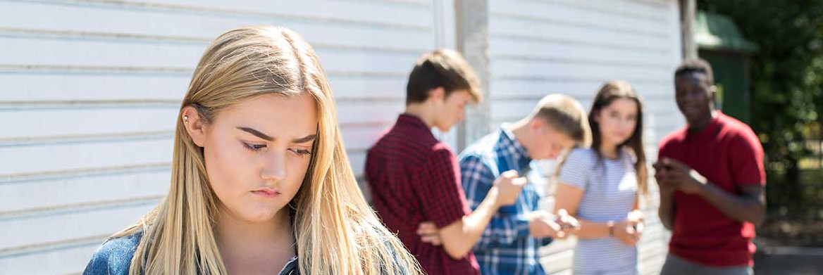 A teenaged girl holding a phone looks sad as three male teens and one female teen stand some distance behind her while looking at their phones or staring at her.
