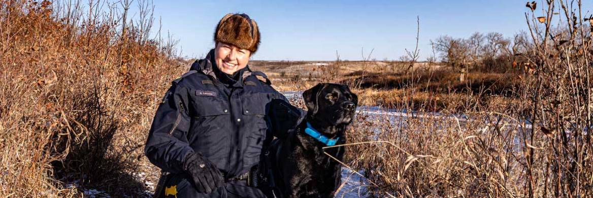 A female police officer in uniform crouches beside a black dog. The officer and dog are in a wooded area in the winter.