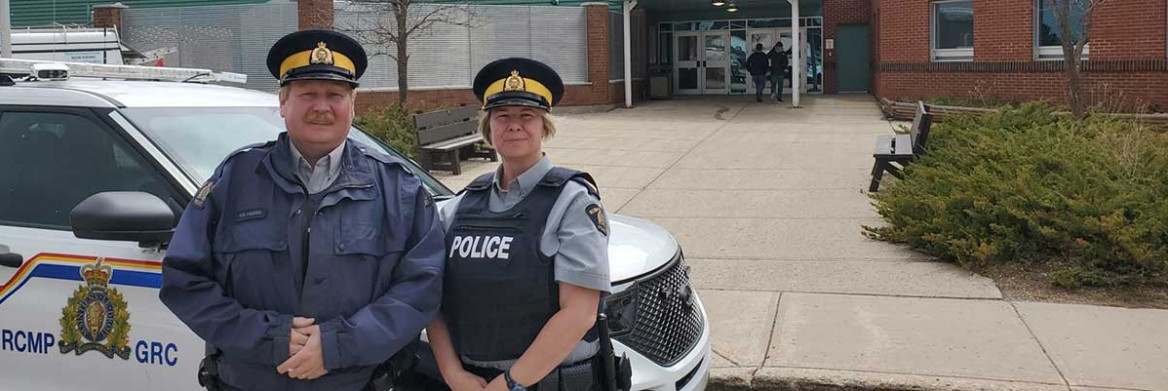 Two RCMP officers stand in front of a police vehicle near a school.