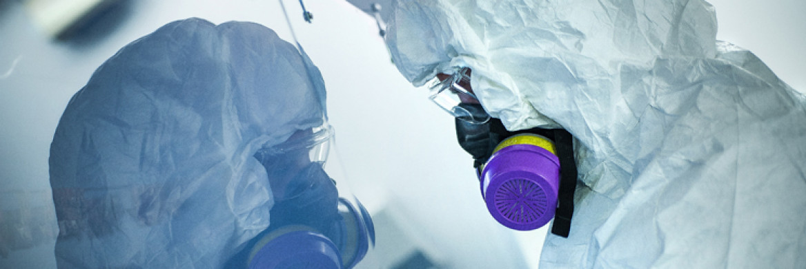 A man wearing protective clothing and a respirator sits in a pristine lab. His reflection appears in a glass wall in front on him.