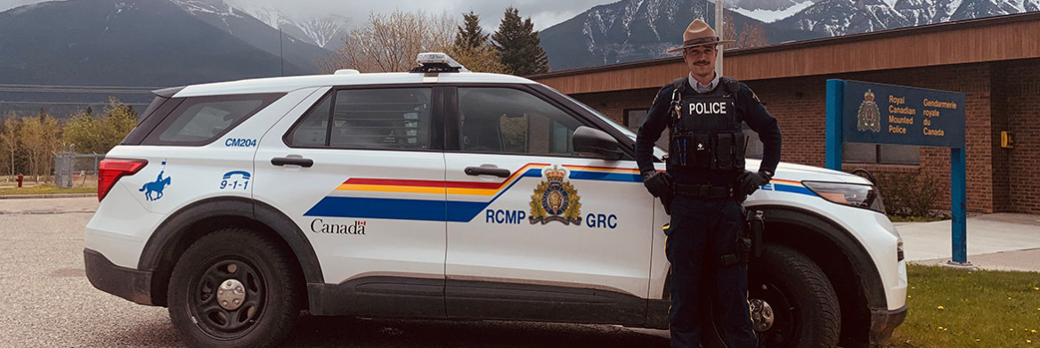 A male RCMP officer stands beside an RCMP hybrid vehicle with the detachment building and cloud-covered mountains in the background.