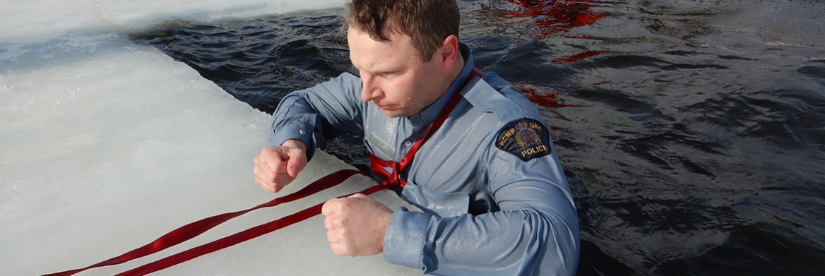 A male RCMP officer stands in icy water with a safety line tied around his chest. 