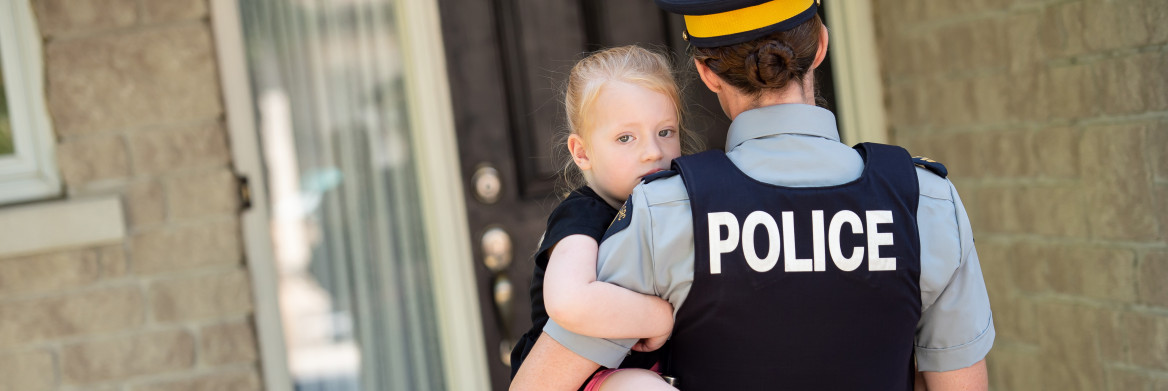 A female police officer holds a young girl in her arms standing at the front door of a house. 