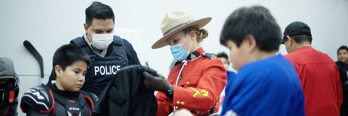An RCMP officer in a regular uniform and an RCMP officer in Red Serge help a boy put on a hockey jersey.
