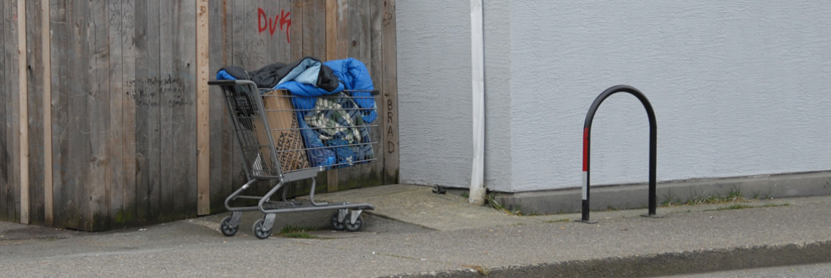 A shopping cart filled with items stands alone on the sidewalk. A fence with grafitti on it is in the background.