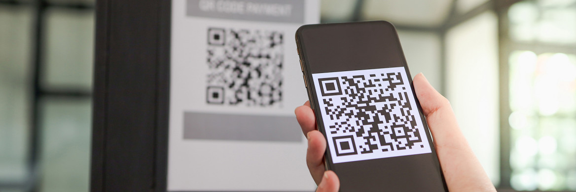 A person's hand holds a smartphone that displays a QR code pattern on its screen. In the background, the same pattern appears on a poster attached to a window.