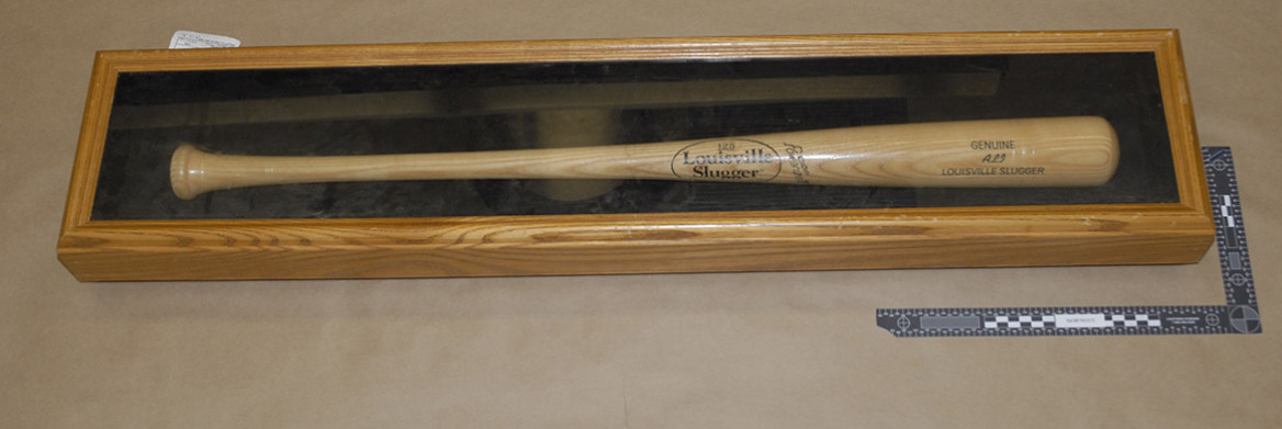 A casefile photo shows a baseball bat encased in a display box. A ruler rests on the right side of the box. 