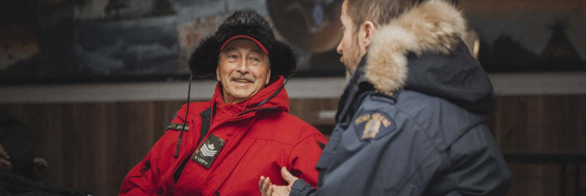  A smiling man wearing a red parka and fur hat sits facing a male RCMP officer wearing a parka.
