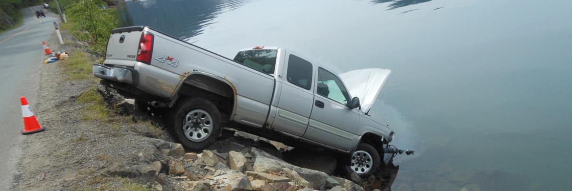 A truck rests on a rocky embankment with its engine hood partially submerged in a lake. Coniferous trees and rugged mountains are in the background.
