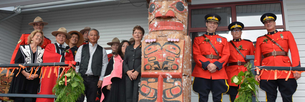 Representatives from the community and the RCMP, including members in red serge, stand next to a totem pole.