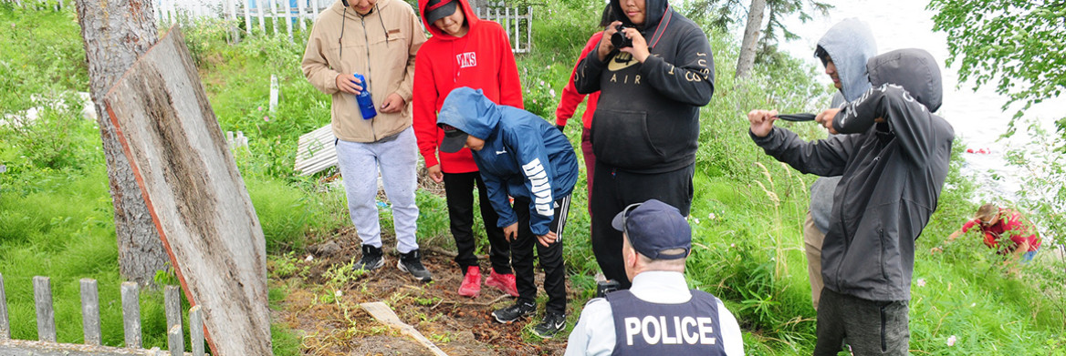 A group of young people and a male RCMP officer look at a patch of soil on the ground.
